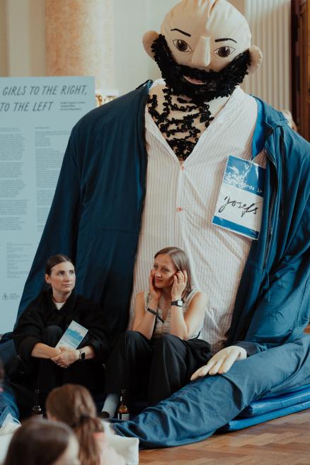 A large textile figure with a beard and two seated women in the foreground, with a display board in the background reading Girls to the Right, to the Left.
