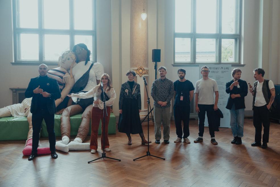 Several people stand in a museum hall by two microphones, with a large soft sculpture installation and tall windows in the background.