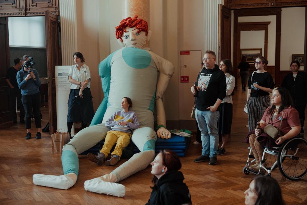 In a museum hall, a large textile sculpture with red curls and blue-and-white limbs sits on the floor, with a woman seated in its lap while visitors stand and sit around it.