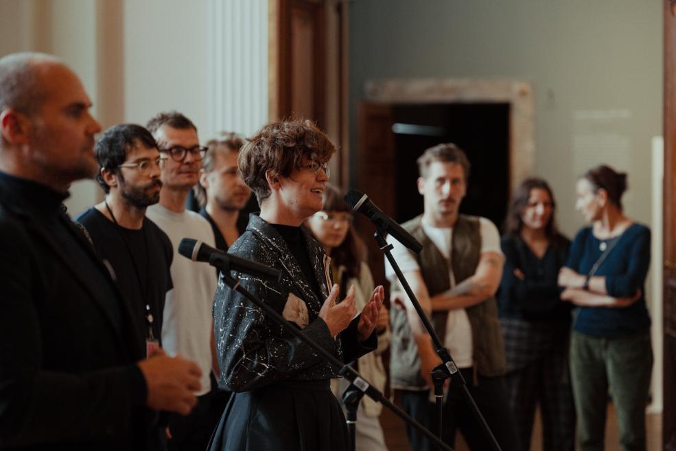 In a museum hall, a short-haired woman wearing glasses speaks at microphones while several listeners stand around her.