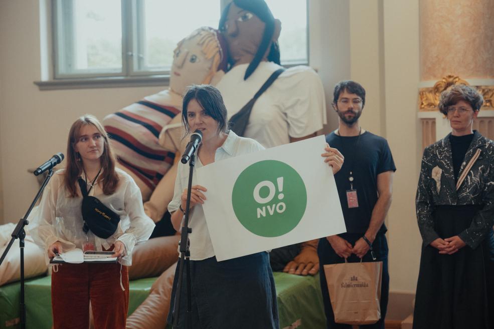 Four people stand by two microphones in a room, and the woman in the center holds a sign with a green circle and the text O! NVO, with a large figurative artwork in the background.
