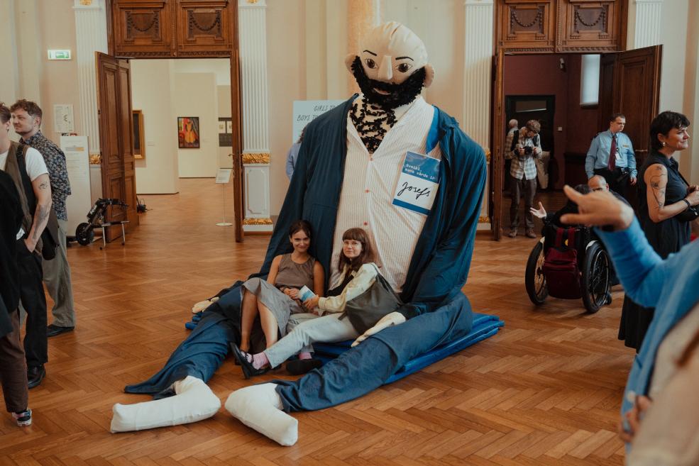 In a museum hall, an installation shows a giant seated figure in blue clothing with two women sitting in its lap, while visitors stand around it.