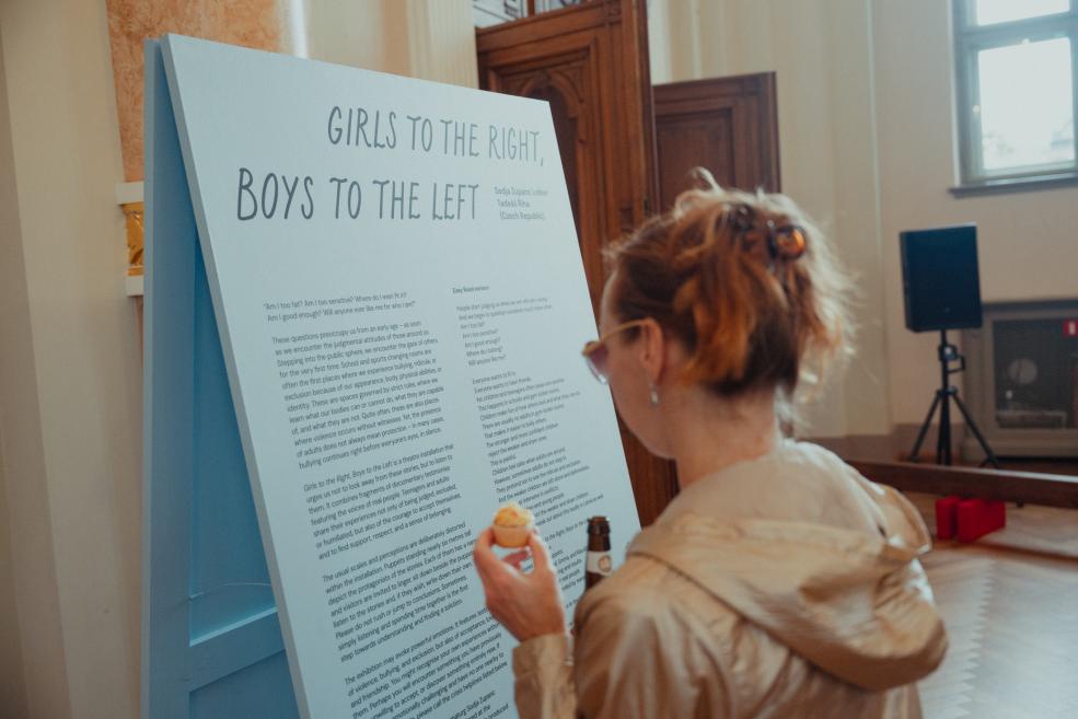 A large blue exhibition display with the title Girls to the Right, Boys to the Left is being viewed by a woman with her hair tied up, holding a small snack in her hand.