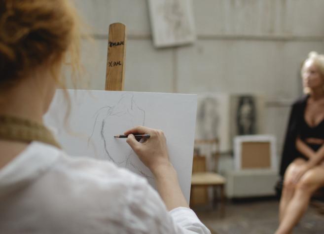 Tēlotājmākslas nodarbība studijā: priekšplānā redzama persona, kas zīmē sēdošu modeli, fonā redzams modelis melnā apģērbā.