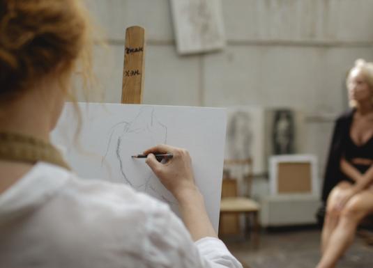 Tēlotājmākslas nodarbība studijā: priekšplānā redzama persona, kas zīmē sēdošu modeli, fonā redzams modelis melnā apģērbā.