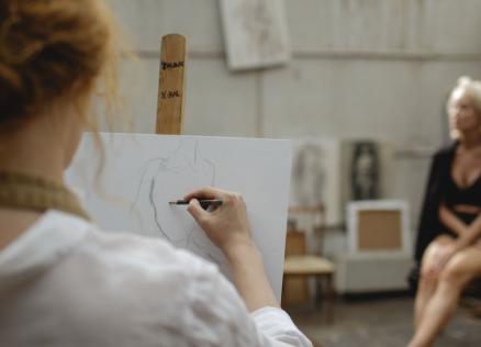 Tēlotājmākslas nodarbība studijā: priekšplānā redzama persona, kas zīmē sēdošu modeli, fonā redzams modelis melnā apģērbā.