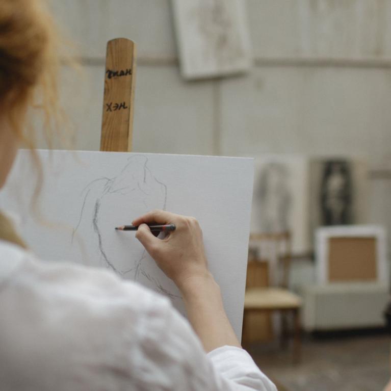 Tēlotājmākslas nodarbība studijā: priekšplānā redzama persona, kas zīmē sēdošu modeli, fonā redzams modelis melnā apģērbā.