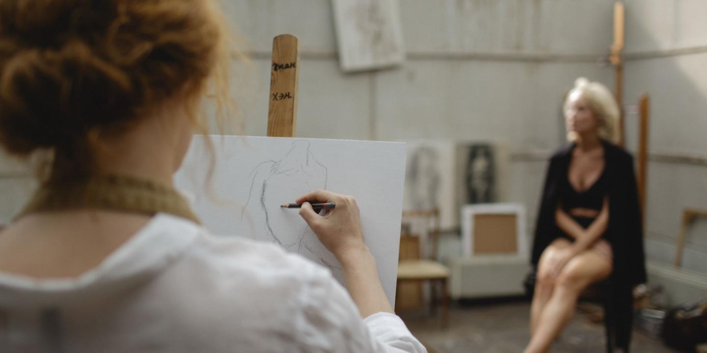 Tēlotājmākslas nodarbība studijā: priekšplānā redzama persona, kas zīmē sēdošu modeli, fonā redzams modelis melnā apģērbā.