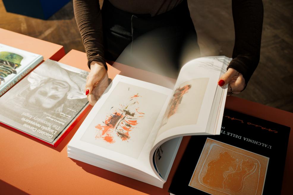 A visitor in an exhibition hall browses an art catalogue on a table with publications, with an artwork reproduction visible on the open page.