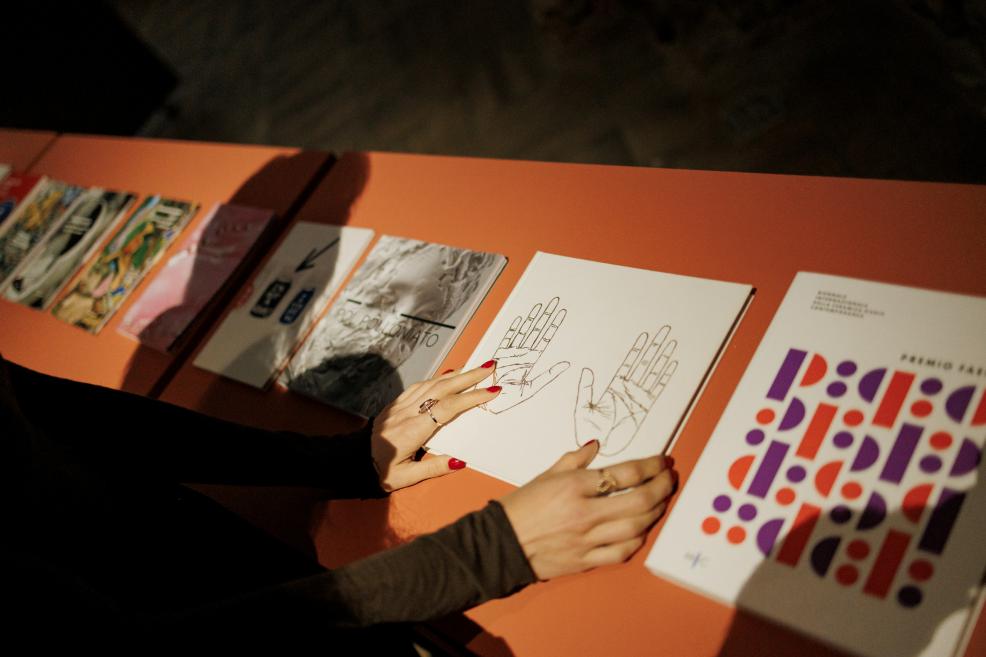 A visitor in a museum exhibition hall examines art catalogues and publications displayed on a table, touching one of the books.