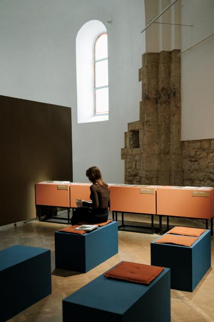 A visitor sits on a bench in a museum exhibition hall reading a publication, surrounded by display cases with printed materials and seating for visitors.