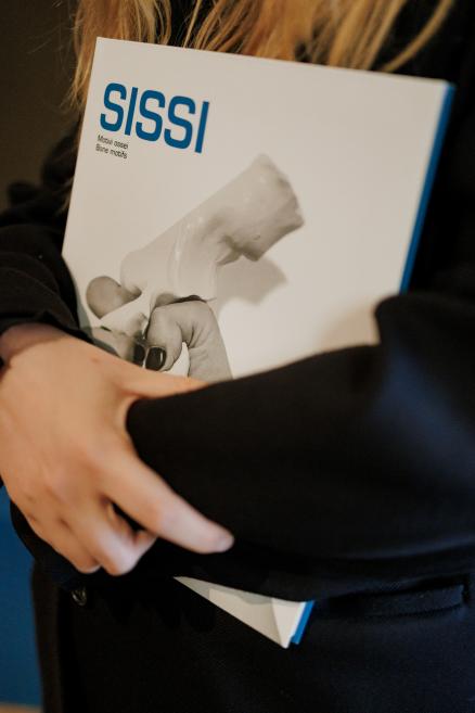 Apmeklētāja muzeja izstāžu zālē rokās tur mākslas katalogu “SISSI. Motel Sissi. Erotic Notes”, kura vāku rotā melnbalta fotogrāfija.