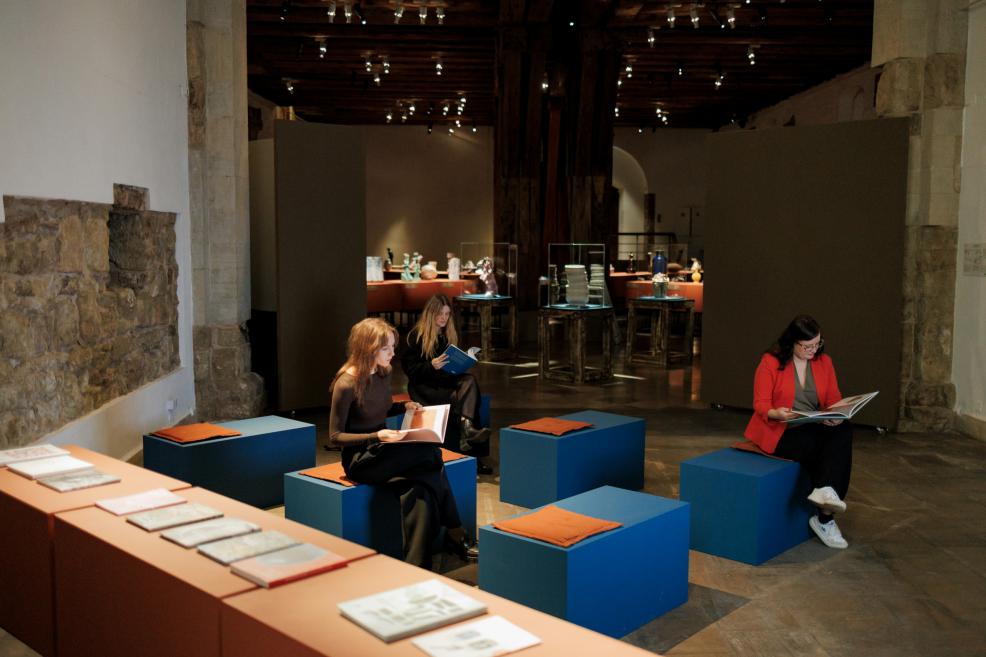 Visitors in a museum exhibition hall sit on benches reading art catalogues, with publications displayed on a table in the foreground and the exhibition installation visible in the background.