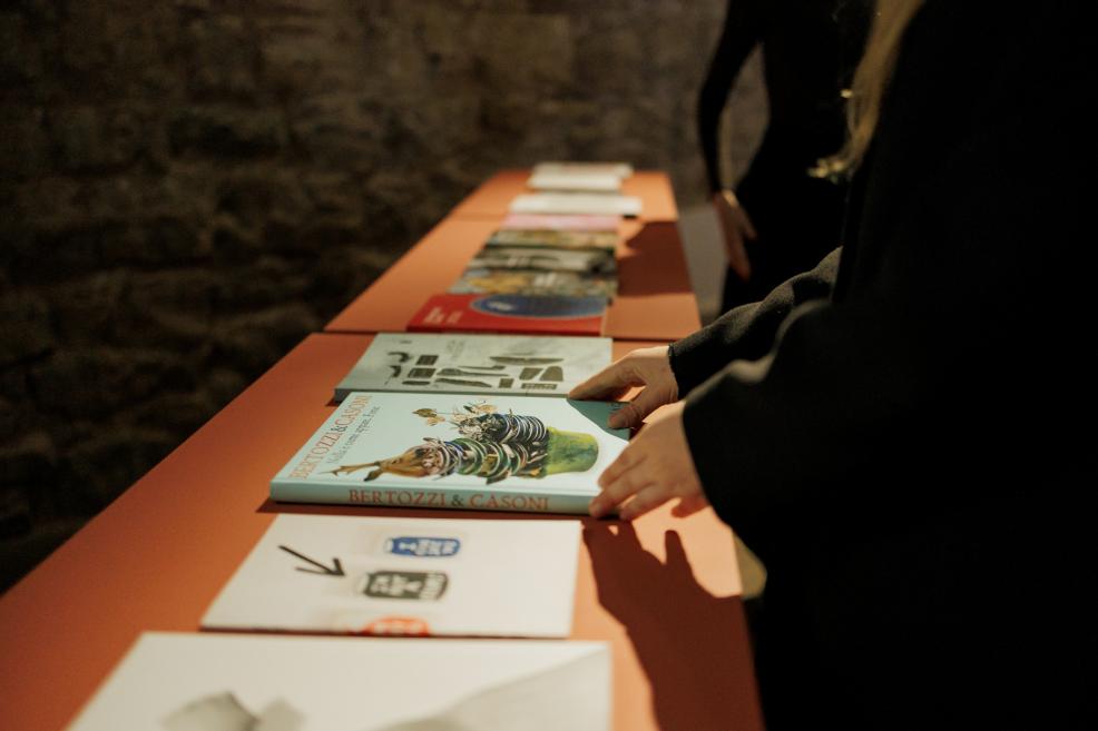 A visitor in a museum exhibition hall examines art catalogues and publications displayed on a table, selecting one of the books.