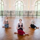 Trīs sievietes sēž meditācijas pozā plašā vēsturiskā zālē ar parketa grīdu un dekoratīvām margām, salikušas plaukstas lūgšanas žestā.