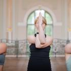 Three women in a bright historic museum hall perform the Eagle Pose arm variation, crossing their arms and pressing their palms together in front of their faces.
