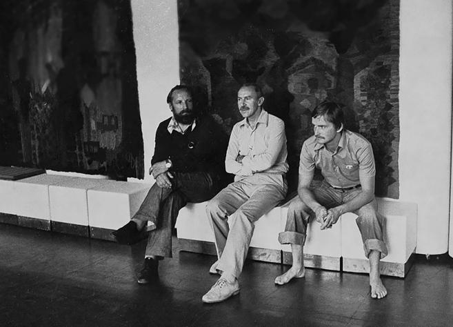 Melnbalta fotogrāfija, kurā trīs vīrieši sēž izstāžu telpā uz soliņa, fonā redzami liela izmēra tekstilmākslas darbi.