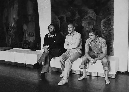 Melnbalta fotogrāfija, kurā trīs vīrieši sēž izstāžu telpā uz soliņa, fonā redzami liela izmēra tekstilmākslas darbi.