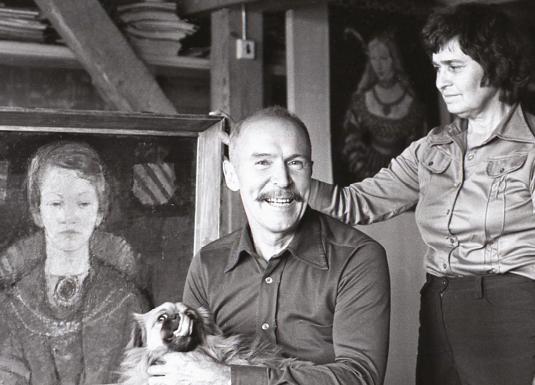 Black-and-white photograph of Ojārs Ābols seated in a studio with a dog on his lap, next to a woman and a framed portrait painting.