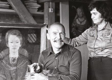 Black-and-white photograph of Ojārs Ābols seated in a studio with a dog on his lap, next to a woman and a framed portrait painting.