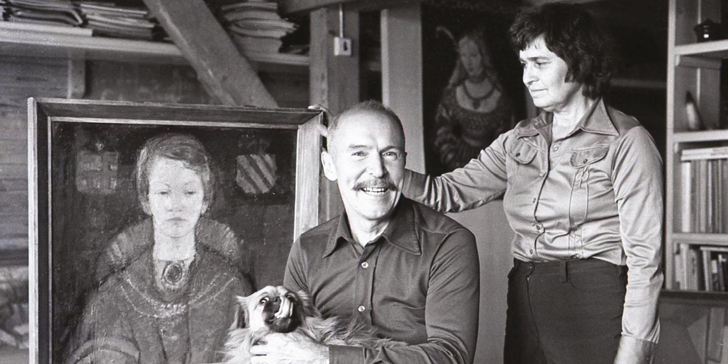 Melnbalta fotogrāfija, kurā redzams mākslinieks ar suni klēpī un sieviete viņam blakus, fonā stāvot gleznai.