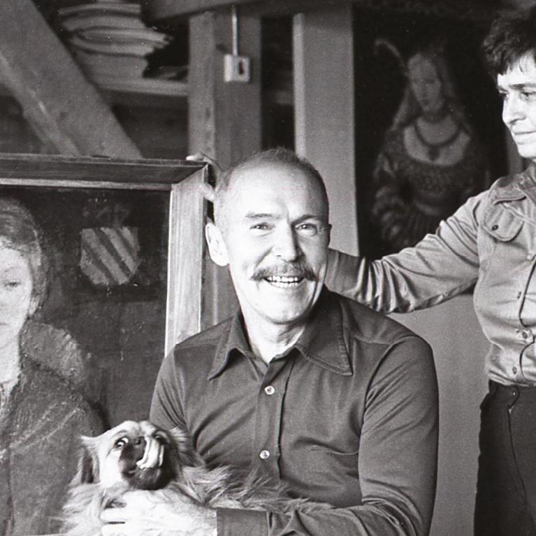 Melnbalta fotogrāfija, kurā redzams mākslinieks ar suni klēpī un sieviete viņam blakus, fonā stāvot gleznai.