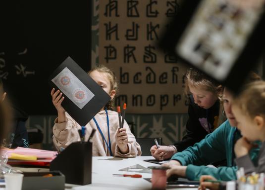 Bērni piedalās muzeja radošajā darbnīcā, zīmējot un krāsojot; viens bērns rokās tur savu darbu ar ornamentālu zīmējumu uz melna papīra.