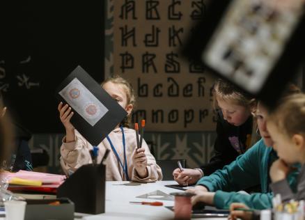 Bērni piedalās muzeja radošajā darbnīcā, zīmējot un krāsojot; viens bērns rokās tur savu darbu ar ornamentālu zīmējumu uz melna papīra.