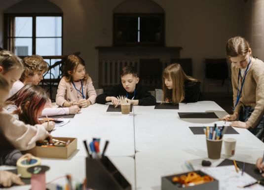 Bērni sēž pie galda radošajā darbnīcā, koncentrējušies uz zīmēšanu un krāsošanu ar dažādiem materiāliem un instrumentiem.