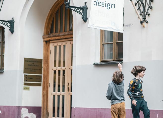 Divi bērni rotaļājas pie Dekoratīvās mākslas un dizaina muzeja ieejas Vecrīgā, pie ēkas ar gaišām sienām un koka durvīm.