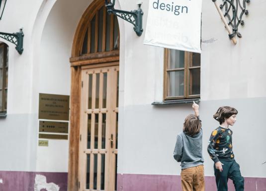 Divi bērni rotaļājas pie Dekoratīvās mākslas un dizaina muzeja ieejas Vecrīgā, pie ēkas ar gaišām sienām un koka durvīm.