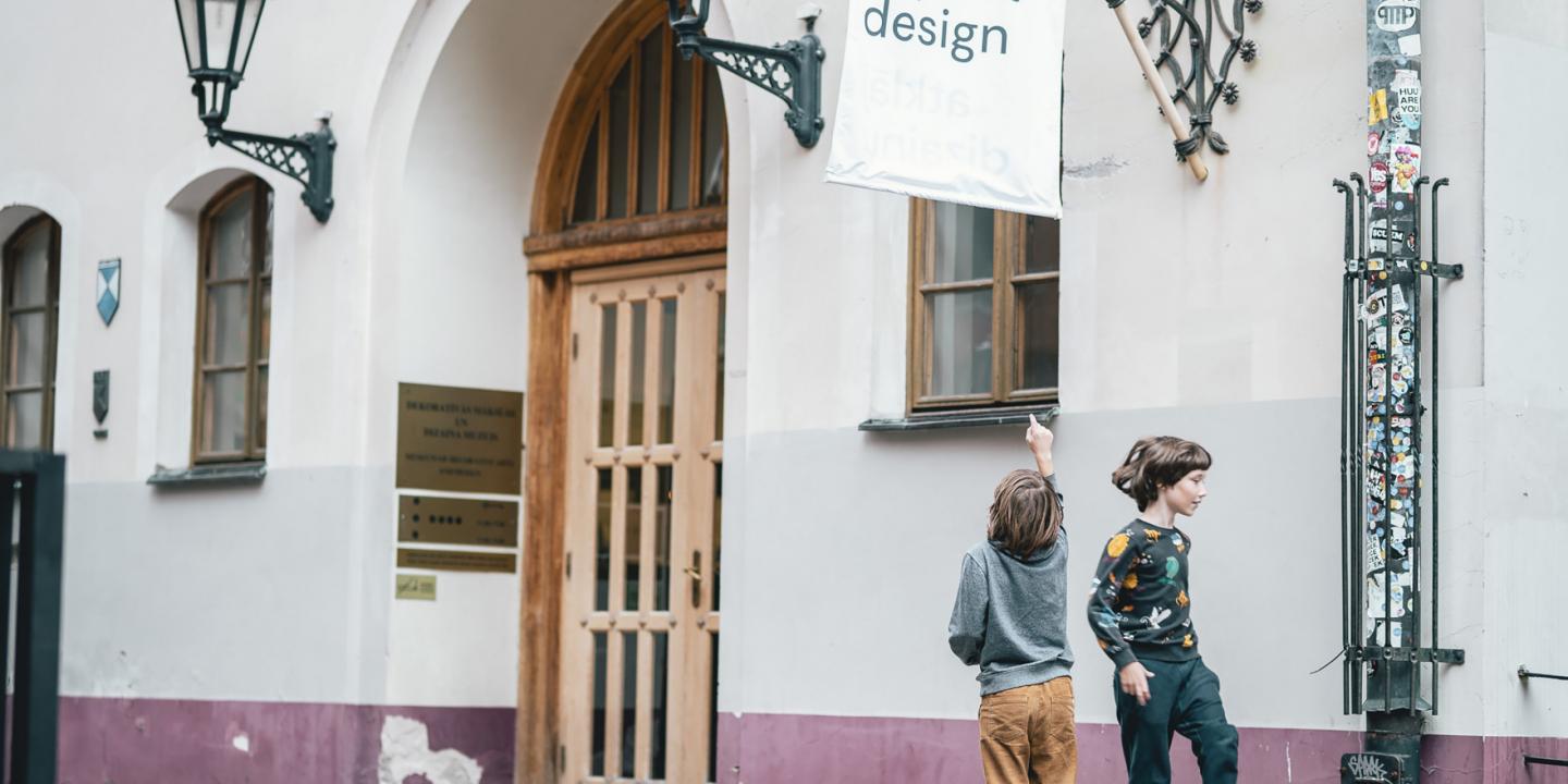 Divi bērni rotaļājas pie Dekoratīvās mākslas un dizaina muzeja ieejas Vecrīgā, pie ēkas ar gaišām sienām un koka durvīm.