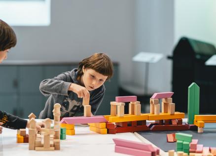 Divi bērni rotaļājas pie Dekoratīvās mākslas un dizaina muzeja ieejas Vecrīgā, pie ēkas ar gaišām sienām un koka durvīm.