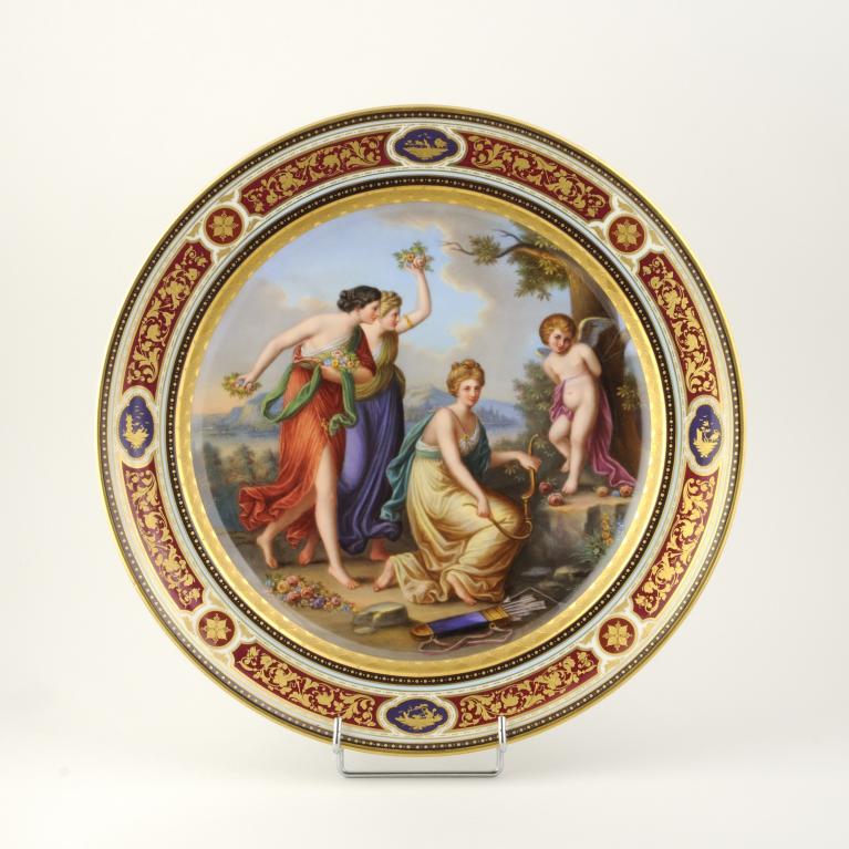 Dekoratīvs porcelāna šķīvis ar klasisku figūru kompozīciju un ornamentālu zeltītu apmali.