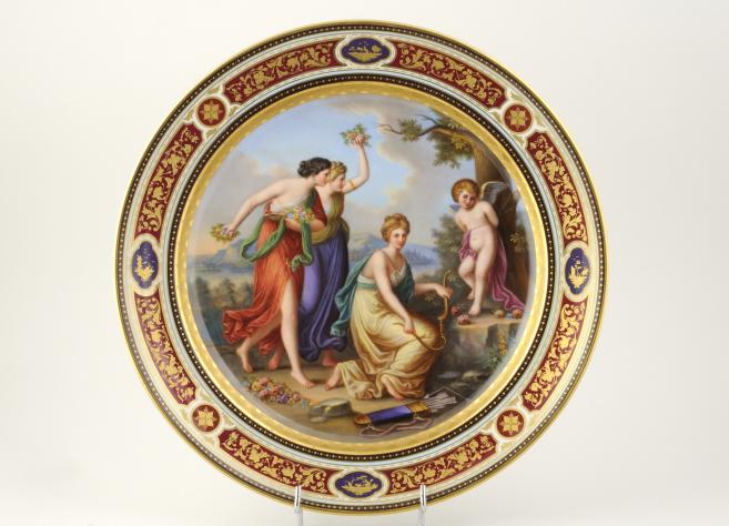 Dekoratīvs porcelāna šķīvis ar klasisku figūru kompozīciju un ornamentālu zeltītu apmali.