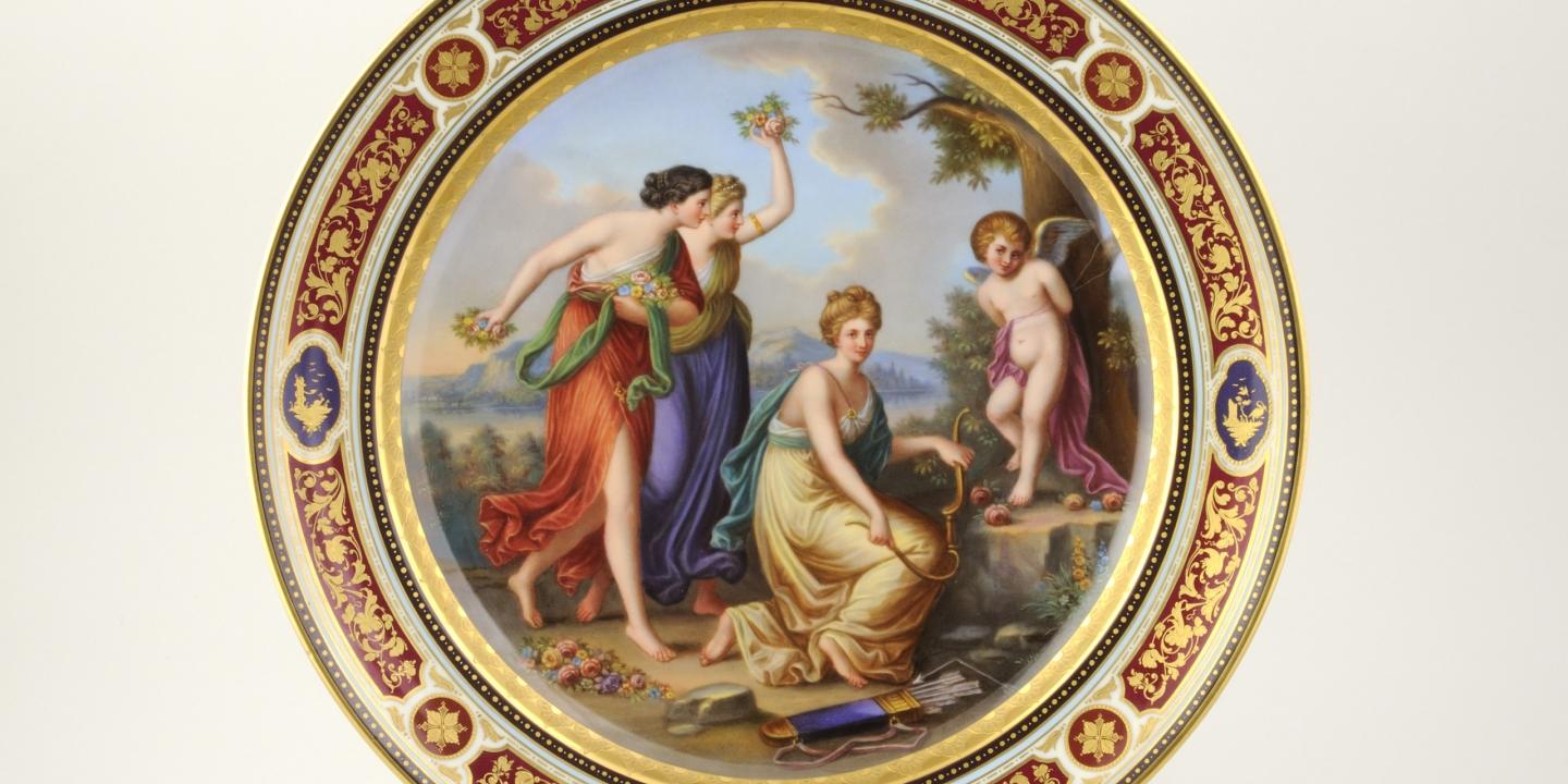 A decorative porcelain plate featuring a classical figural scene and an ornate gilded border.