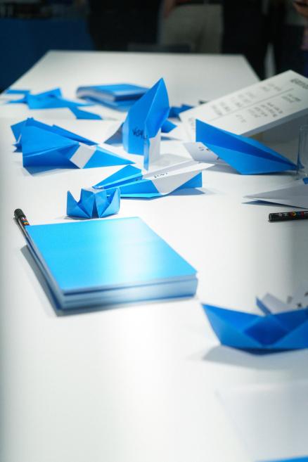 Uz balta galda izvietotas zilas un baltas papīra origami figūras, piezīmju blociņš un marķieri.