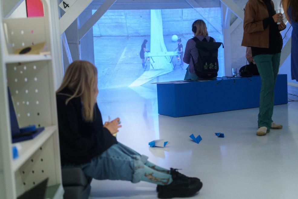 Galerijas telpā vairāki apmeklētāji skatās zilu video projekciju uz sienas, sēžot un stāvot pie zilas platformas un baltām plauktu konstrukcijām.