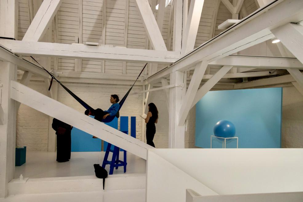 Baltā galerijas telpā ar koka sijām divi cilvēki stāv pie zila instalācijas darba ar piekārtu šūpuļtīklu un lielu zilu sfēru uz pjedestāla.