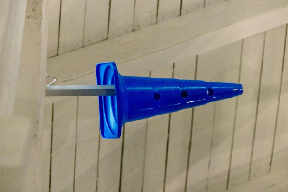 Zilā plastmasas konusa formas detaļa uz metāla stieņa uz gaiša koka fona.