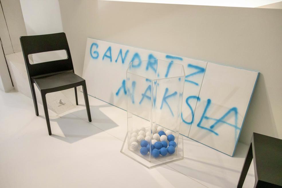 Galerijas instalācija ar melnu krēslu, caurspīdīgu kasti ar baltām un zilām bumbiņām un baltiem paneļiem ar zilu grafiti uzrakstu.