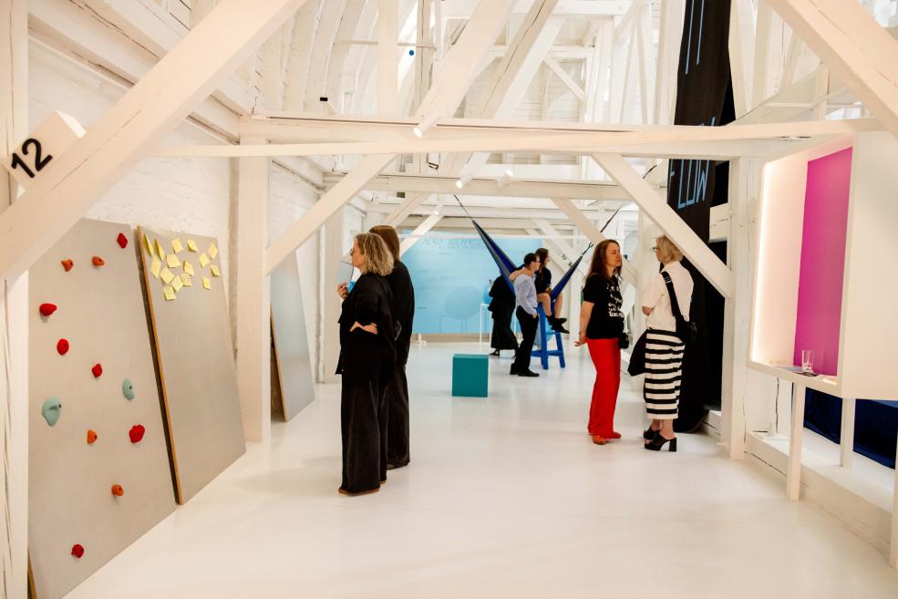 Baltā galerijas telpā ar koka sijām apmeklētāji skatās instalācijas ar krāsainām piezīmēm, paneļiem un šūpuļtīklu.