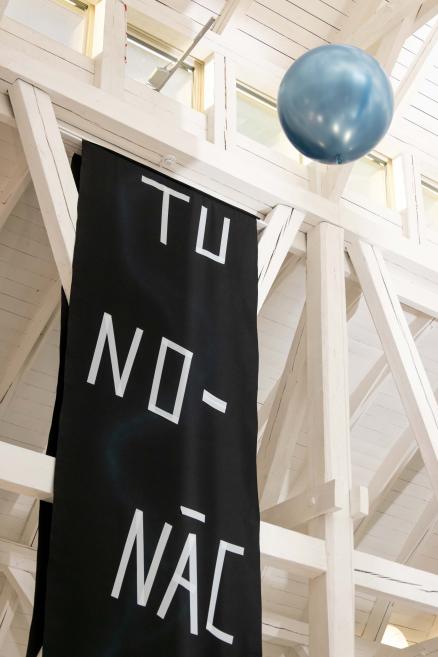 Melns vertikāls baneris ar baltām burtiem pie balti krāsotas koka konstrukcijas, blakus zils balons interjerā.