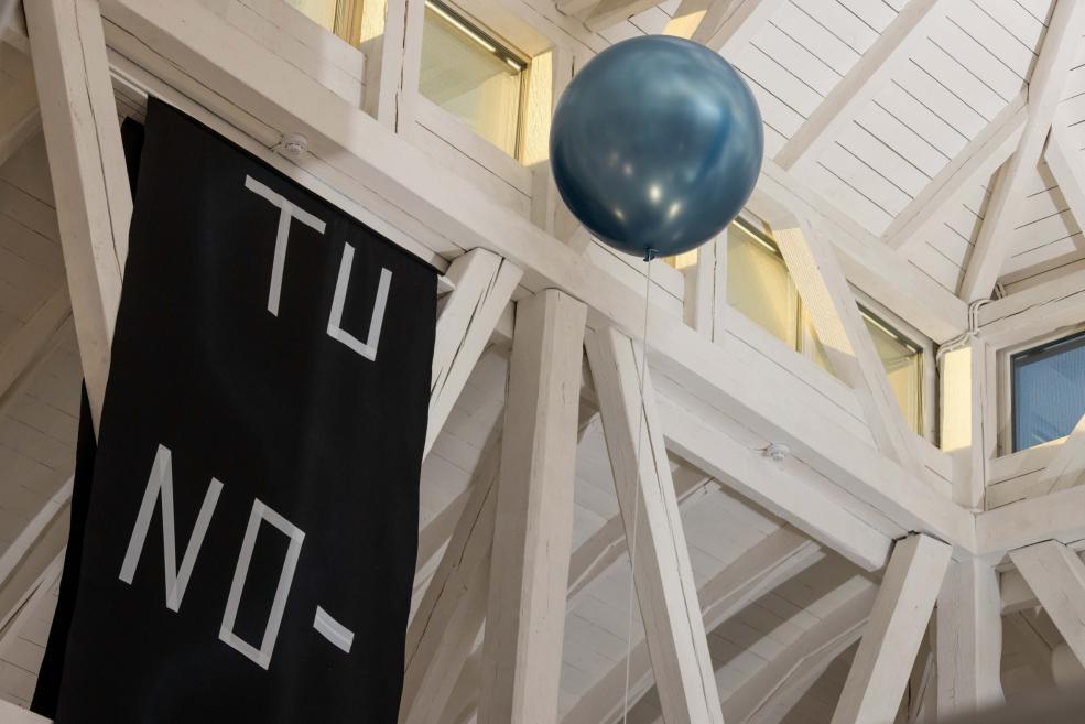 Muzeja interjerā pie baltiem koka sijām karājas zils balons un melns baneris ar baltu uzrakstu TU NO-.