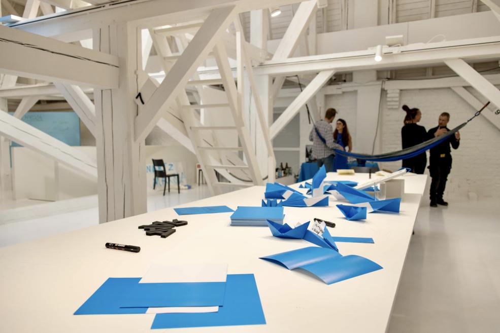 Baltā galerijas telpā pie gara galda ar zilām papīra maketiem un marķieriem stāv trīs cilvēki, fonā redzamas baltas koka konstrukcijas un piekārts audums.