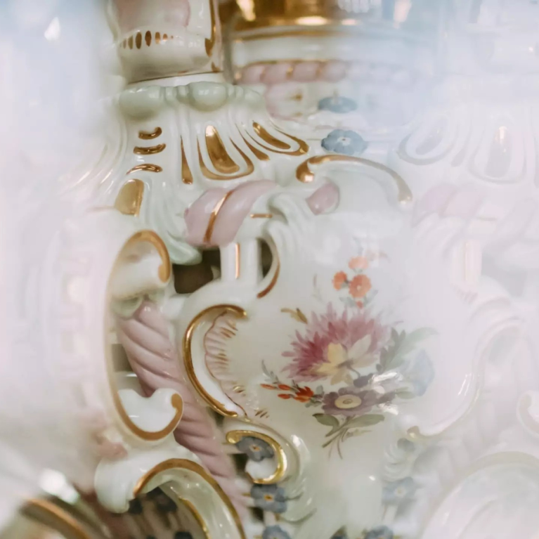 Close-up of a white porcelain decorative detail with gilding, pastel-coloured ornamentation and a painted floral composition.