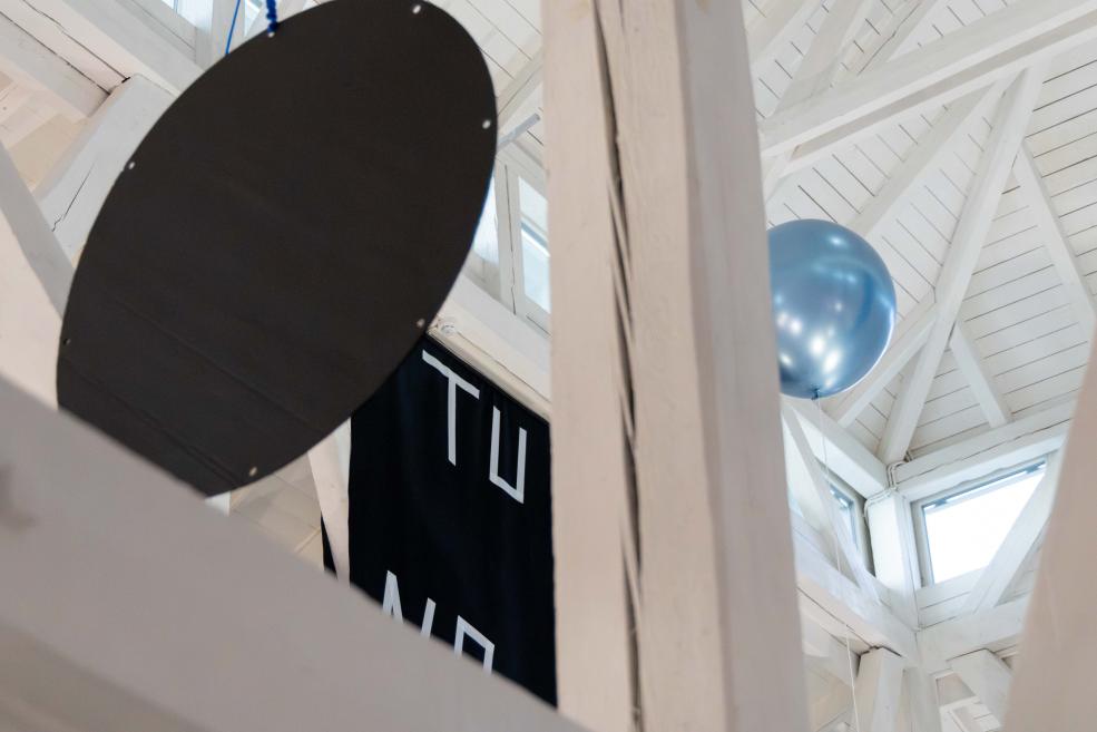 Balta koka interjerā redzami liels melns ovāls elements, zils balons un melns tekstila baneris ar baltām burtnēm.
