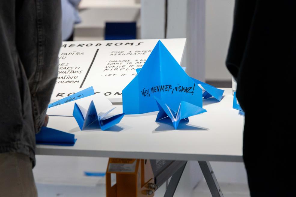 Uz balta galda izlikti zilas papīra origami lidmašīnas un plakāts ar tekstu Aerodroms.