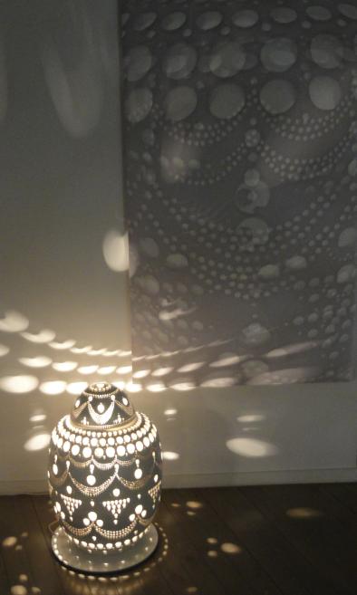 Tumša, ar apaļiem caurumiem rotāta dekoratīva lampa uz grīdas, kas met gaismas punktu rakstus uz sienas un grīdas.