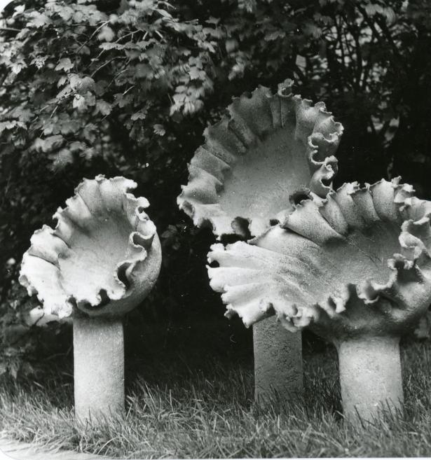Melnbalta fotogrāfija ar trim viļņainām skulpturālām formām uz cilindriskiem balstiem zālē krūmu fonā.